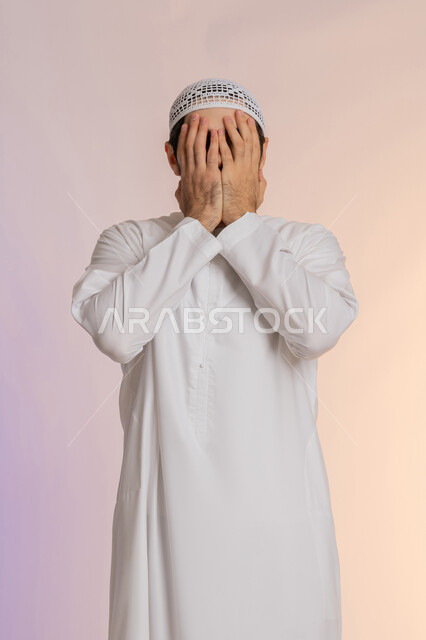 الشعور بالخجل والإحراج، إغماض العينين وعدم الرغبة بالرؤية، البكاء ومشاعر الحزن، التعرض لصدمة وعدم التصديق، بورتريه لرجل عربي خليجي اماراتي يرتدي الكندورة و القبعة يغطي وجهه بيديه، خلفية ملونة