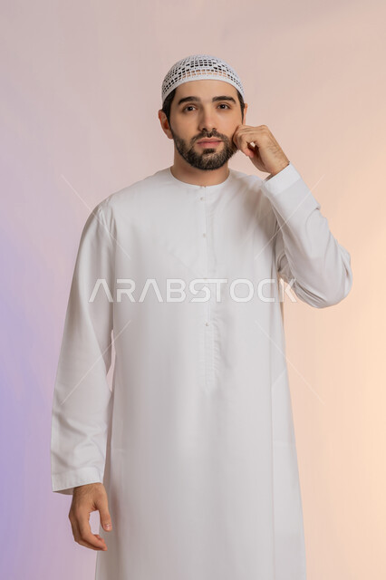 Caring about one's appearance, the concept of handsomeness and self-confidence, standing and looking at the camera with gestures of pleasure and cheerfulness, a portrait of an Arab Gulf Emirati man wearing a kandura and a white hat, standing and putting his hand on his cheek, authentic Arab manhood, colorful background