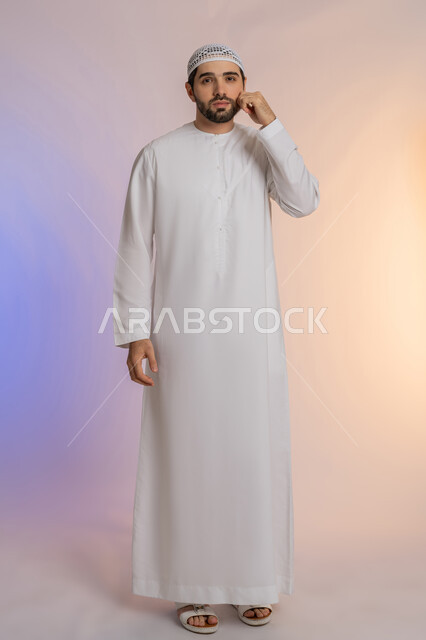مفهوم الوسامة والثقة بالنفس، الوقوف والنظر إلى الكاميرا بإيماءات السرور والبشاشة، بورتريه لرجل عربي خليجي اماراتي يرتدي الكندورة والقبعة البيضاء يقف ويضع يده على خده، الرجولة العربية الأصيلة، الاهتمام بالمظهر الخارجي، الجسم بالطول الكامل، خلفية ملونة