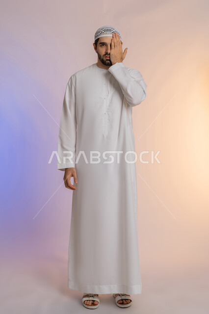 Placing the hand on the eye and covering it, inflammation and redness of the eyes and the need to visit a doctor, double vision and blurred vision, portrait of an Arab Gulf Emirati man wearing a kandora and a hat suffering from health problems in his eyes, full length body, colored background.