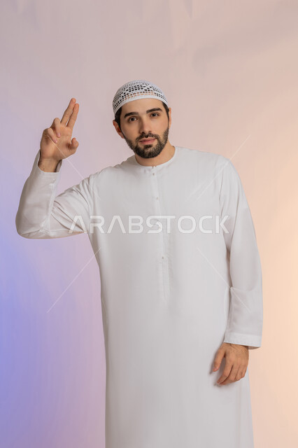 مفهوم التقدير والاحترام، الأناقة والوسامة والرجولة، إلقاء التحية والسلام، بورتريه لرجل عربي خليجي اماراتي مبتسم يرتدي الكندورة والقبعة  ينظر إلى الكاميرا بإيماءات السعادة والسرور، الوقوف باستقامة بتعابير الثقة بالنفس، خلفية ملونة