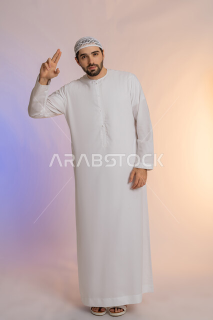 إلقاء التحية والسلام، الأناقة والوسامة والرجولة، مفهوم التقدير والاحترام، بورتريه لرجل عربي خليجي اماراتي مبتسم يرتدي الكندورة والقبعة  ينظر إلى الكاميرا بإيماءات السعادة والسرور، الوقوف باستقامة بتعابير الثقة بالنفس، الجسم بالطول الكامل، خلفية ملونة
