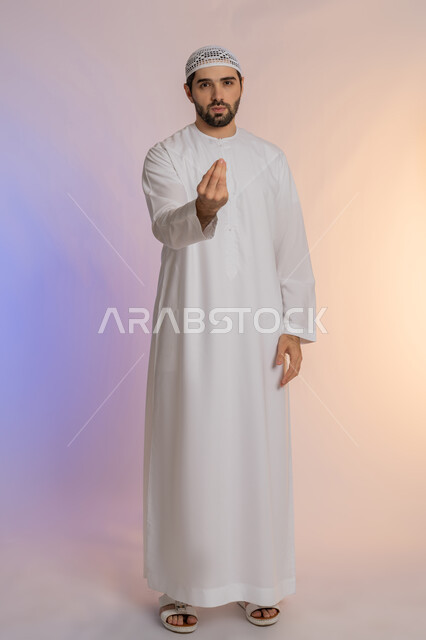 مفهوم الصبر والتريث و التأني, التهديد و التوعد لشيء ما، بورتريه لرجل عربي خليجي اماراتي  يرتدي الكندورة والقبعة ينظر الى الكاميرا يرفع يده نحو الأعلى بإيماءات طلب الإنتظار, الجسم بالطول الكامل، خلفية ملونة
