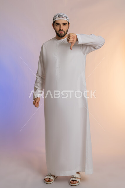 الإشارة باصبع الإبهام نحو الأسفل بعلامات السوء وعدم القبول، الشعور بالانزعاج والاضطراب، التعبير عن الجودة السيئة، بورتريه لرجل عربي خليجي اماراتي يرتدي الكندورة والقبعة  ينظر للكاميرا بإيماءات الغضب، الجسم بالطول الكامل، خلفية ملونة