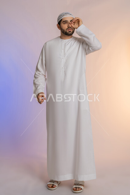 التخطيط للأهداف والطموحات المستقبلية، محاولة تقريب الأشياء البعيدة، التركيز على شيء ما، بورتريه لرجل عربي خليجي اماراتي يرتدي الكندورة والقبعة يضع يده على عينه بشكل عدسة بإيماءات البحث، الجسم بالطول الكامل، خلفية بيضاء