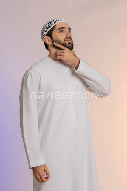 The concept of listening and focusing, solving problems and making decisions, looking forward and planning for the future, portrait of an Arab Gulf Emirati man wearing a kandora and a hat, placing his hand on his chin with gestures of thought and absent-mindedness, a colored background.