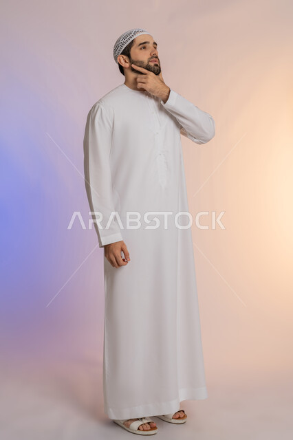 حل المشكلات واتخاذ القرارات، التطلع والتخطيط للمستقبل، بورتريه لرجل عربي خليجي اماراتي يرتدي الكندورة والقبعة يضع يده على ذقنه بإيماءات التفكير وشرود الذهن، مفهوم الاستماع والتركيز، صورة الجسم بالطول الكامل، خلفية ملونة