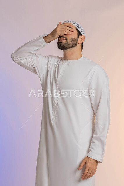 حركات اليدين والوجه تعبر عن الخجل، ايماءات تذكر شيء ما بشكل مفاجئ، بورتريه لرجل عربي خليجي اماراتي يرتدي الكندورة والقبعة يقف بشكل مستقيم ويغطي عينيه بيده، الاهتمام بالمظهر الخارجي، خلفية ملونة