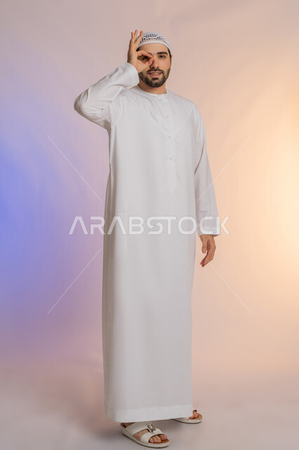 محاولة تقريب الأشياء البعيدة، التخطيط للأهداف والطموحات المستقبلية، حب الاكتشاف والمعرفة والمغامرة، التركيز على شيء ما، بورتريه لرجل عربي خليجي اماراتي يرتدي القبعة يضع يده على عينه بشكل عدسة بإيماءات البحث، صورة لكامل الجسم، خلفية ملونة