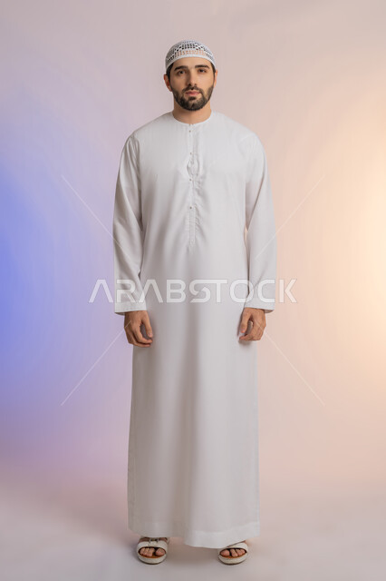 Looking at the camera with gestures of self-confidence, paying attention to external appearance, the concept of elegance and masculinity, a portrait of an Emirati Gulf Arab man wearing a kandora and wearing a hat, standing upright and moderate, taking personal photos for official documents, full-length body, colored background