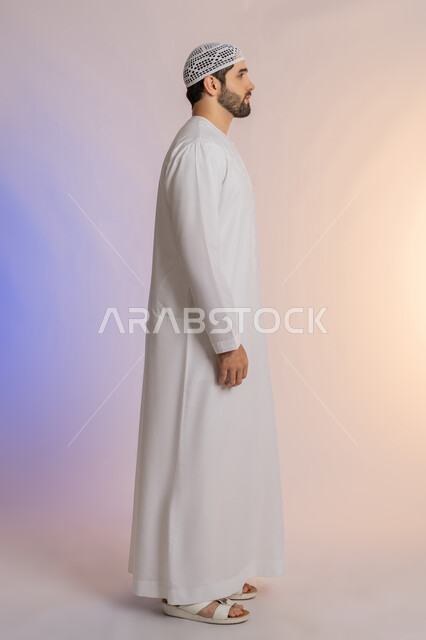 النظر الى مكان ما بايماءات الثقة بالنفس، الاهتمام بالمظهر الخارجي, مفهوم الأناقة والرجولة, بورتريه من الجانب لرجل عربي خليجي اماراتي يرتدي الكندورة والقبعة يقف باستقامة واعتدال, الجسم بالطول الكامل, خلفية ملونة