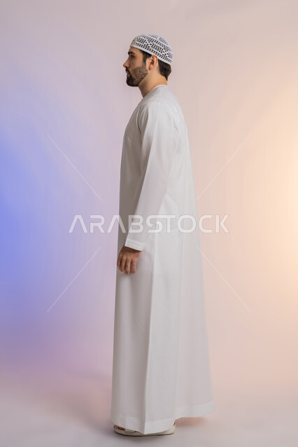 النظر الى مكان ما بايماءات الثقة بالنفس، لاهتمام بالمظهر الخارجي, مفهوم الأناقة والرجولة, بورتريه من الجانب لرجل عربي خليجي اماراتي يرتدي الكندورة والقبعة يقف باستقامة واعتدال, الجسم بالطول الكامل, خلفية ملونة