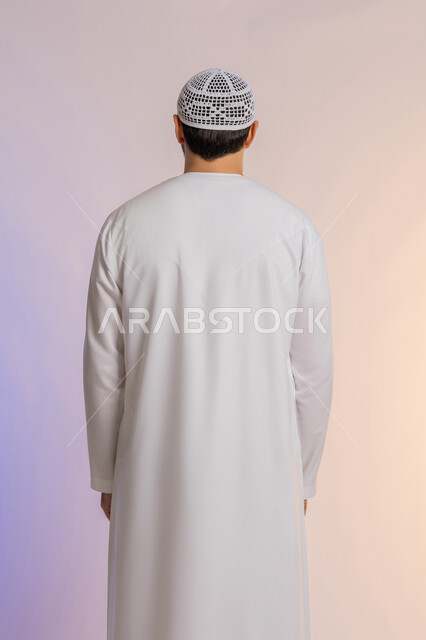 الاهتمام بالمظهر الخارجي, مفهوم الأناقة والرجولة, بورتريه من الخلف لرجل عربي خليجي اماراتي يرتدي الكندورة ويضع القبعة يقف باستقامة واعتدال, خلفية ملونة