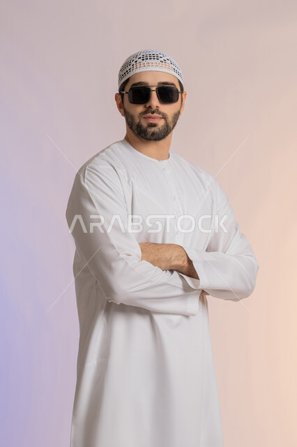 استخدام العدسات الطبية لحماية العين من أشعة الشمس، الأناقة والوسامة والاهتمام بالمظهر الخارجي، بورتريه لرجل عربي خليجي اماراتي  يرتدي الكندورة والطاقية يقف بايدي متقاطعة بإيماءات الثقة بالنفس ويضع نظارة شمسية سوداء، خلفية ملونة