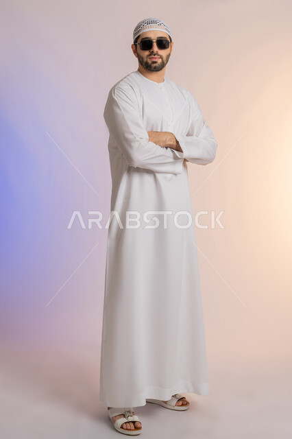 الأناقة والوسامة والاهتمام بالمظهر الخارجي، استخدام العدسات الطبية لحماية العين من أشعة الشمس، بورتريه لرجل عربي خليجي اماراتي  يرتدي الكندورة والطاقية يقف بايدي متقاطعة بإيماءات الثقة بالنفس ويضع نظارة شمسية سوداء، الجسم بالطول الكامل, خلفية ملونة