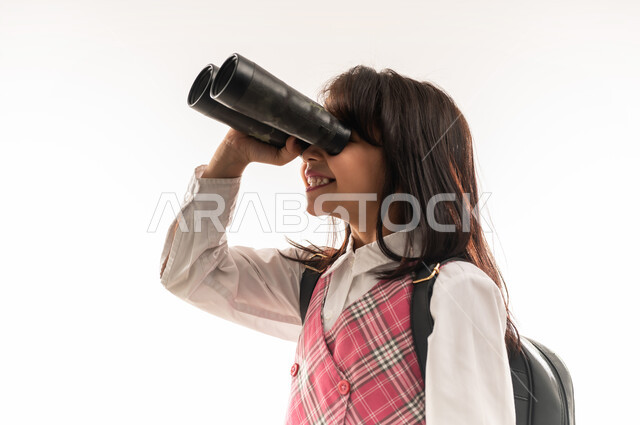 طالبة عمانية ترتدي الزي المدرسي والحقيبة المدرسية تقوم باستخدام المنظار ثنائي العينين لتقريب الاشياء البعيدة، رؤية جديدة لتحسين جودة التعليم في المملكة، بورتريه مقرب من الجانب لتلميذة عربية خليجية سعودية تستعد للفصل الدراسي الجديد، خلفية بيضاء