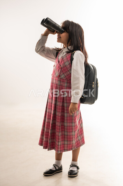 رؤية جديدة لتحسين جودة التعليم في المملكة، طالبة عمانية ترتدي الزي المدرسي والحقيبة المدرسية تقوم باستخدام المنظار ثنائي العينين لتقريب الاشياء البعيدة، بورتريه من الجانب لتلميذة عربية خليجية سعودية تستعد للفصل الدراسي الجديد، صورة الجسم بالطول الكامل، خلفية بيضاء