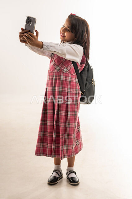 Documenting happy moments, portrait of a Saudi Arabian Gulf student wearing school uniform and school bag taking a commemorative selfie, an Omani student using a mobile phone with gestures of joy and happiness, full body, white background