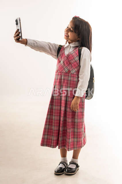 طالبة عمانية تقوم باستخدام الهاتف المحمول بإيماءات الفرح والسرور، مفهوم توثيق اللحظات السعيدة، بورتريه لتلميذة عربية خليجية سعودية ترتدي الزي المدرسي والحقيبة المدرسية تقوم بالتقاط صورة السيلفي التذكارية، الجسم بالطول الكامل، خلفية بيضاء