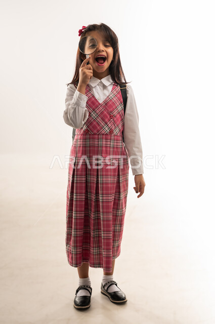 المساعدة على الاستكشاف والبحث، بورتريه لتلميذة عربية خليجية سعودية ترتدي الزي المدرسي والحقيبة المدرسية تنظر عبر العدسة المكبرة، طالبة عمانية تستعد للفصل الدراسي الجديد، مفهوم التدقيق والتركيز وتوضيح الصورة، الجسم بالطول الكامل، خلفية بيضاء