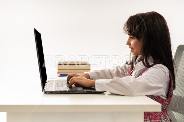 The concept of distance learning, following online classes via the Internet, a close-up portrait of a Saudi Arabian Gulf student wearing a school uniform working on a laptop, using modern technology, an Omani student sitting at a study table with an expression of pleasure, white background