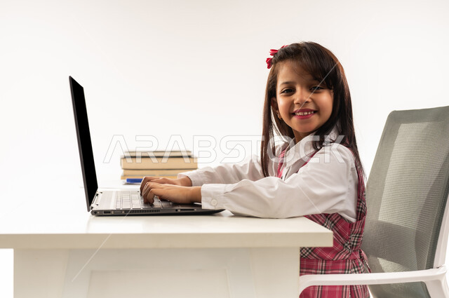 متابعة حصص الاونلاين عن طريق جهاز الحاسوب المحمول, بورتريه  مقرب من الجانب لطالبة عربية خليجية سعودية ترتدي اليونيفووم المدرسي تنظر الى الكاميرا بايماءات الفرح، تلميذة عمانية تجلس على طاولة الدراسة تقوم باستخدام التكنولوجيا الحديثة المتطورة، خلفية بيضاء