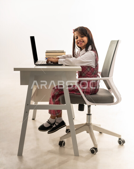 مفهوم التعلم عن بعد باستخدام التكنولوجيا الحديثة، بورتريه من الجانب لطالبة عربية خليجية سعودية ترتدي اليونيفووم المدرسي تنظر الى الكاميرا بايماءات الفرح، تلميذة عمانية تجلس على طاولة الدراسة تقوم بمتابعة حصص الاونلاين عن طريق جهاز الحاسوب المحمول، خلفية بيضاء