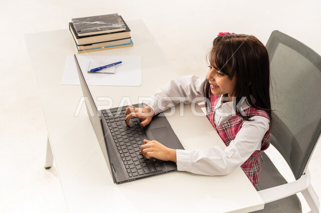 Follow online classes via the internet, portrait from above of a Saudi Arabian Gulf student wearing a school uniform working on a laptop, the concept of distance learning using modern technology, an Omani student sitting at a study table with an expression of pleasure, white background