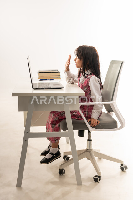 Following lessons and assignments through distance learning platforms, active student participation, side portrait of a Saudi Arabian Gulf student wearing school uniform attending her lessons via laptop, Omani student sitting at study table raising her hand to answer, white background