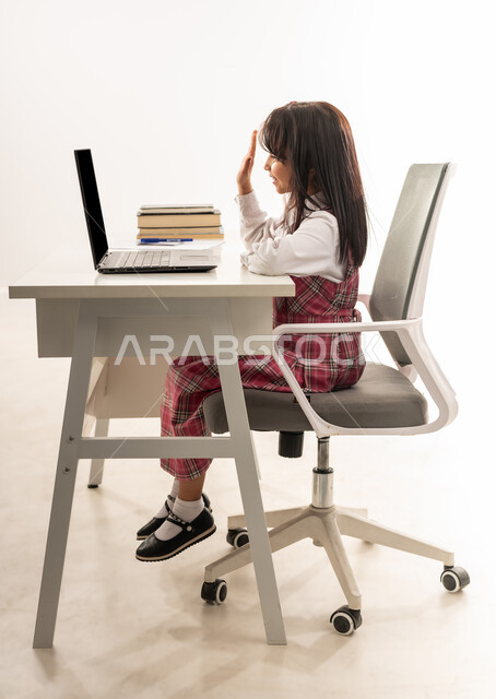 المشاركة الفعالة للطلاب، متابعة الدروس والواجبات من خلال منصات التعلم عن بعد، بورتريه من الجانب لتلميذة عربية خليجية سعودية ترتدي الزي المدرسي تحضر دروسها عن طريق جهاز الحاسوب المحمول، طالبة عمانية تجلس على طاولة الدارسة ترفع يدها للإجابة، خلفية بيضاء