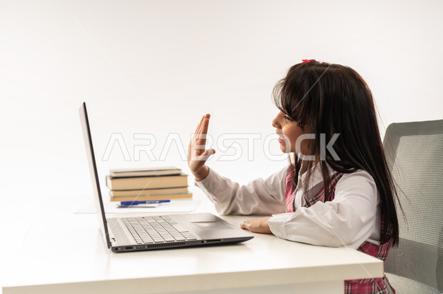 Following lessons and assignments through distance learning platforms, close-up portrait from the side of a Saudi Arabian Gulf student wearing school uniform attending her lessons via laptop, an Omani student sitting at a study table raising her hand to answer, active student participation, white background