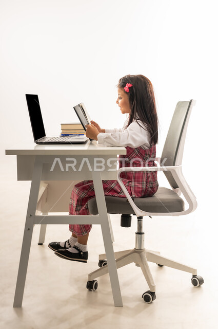متابعة الدروس والواجبات من خلال منصات التعلم عن بعد، المشاركة الفعالة للطلاب، بورتريه لتلميذة عربية خليجية سعودية ترتدي الزي المدرسي تحضر دروسها عن طريق جهاز الحاسوب، طالبة عمانية تجلس على طاولة الدارسة تحمل الكتاب للتأكد من الإجابة، خلفية بيضاء