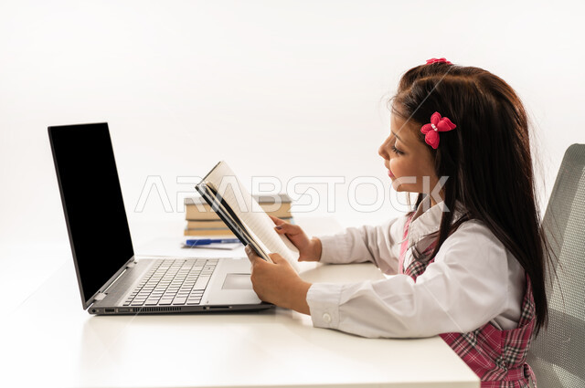 المشاركة الفعالة للطلاب، متابعة الدروس والواجبات من خلال منصات التعلم عن بعد، بورتريه مقرب لتلميذة عربية خليجية سعودية ترتدي الزي المدرسي تحضر دروسها عن طريق جهاز الحاسوب، طالبة عمانية تجلس على طاولة الدارسة تحمل الكتاب للتأكد من الإجابة، خلفية بيضاء