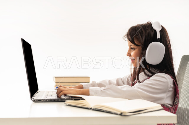 E-learning concept, distance learning using technology, close-up side portrait of a Saudi Arabian Gulf female student wearing school uniform and headphones following lessons using computer, Omani female student sitting at study table with happy expression, white background