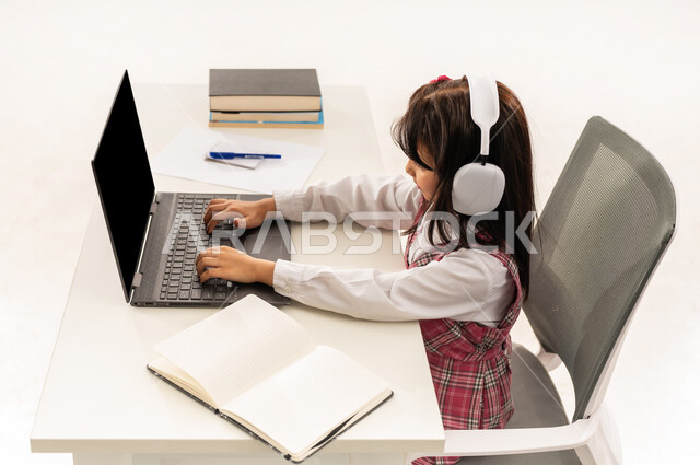 E-learning concept, distance learning using modern technology, portrait of a Saudi Arabian Gulf student wearing school uniform and headphones following lessons using computer, Omani student sitting at study table with happy expressions, white background