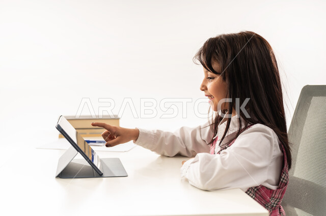 استخدام الاجهزة التقنية الحديثة، تلميذة سعودية تتابع الحصص الدرسية تقوم بحل الواجبات عبر الانترنت، بورتريه مقرب من الجانب لفتاة عربية خليجية عمانية ترتدي الزي المدرسي تقوم بمراجعة دروسها عبر جهاز التابلت، مفهوم الدراسة عن بعد و التعليم الإلكتروني، خلفية بيضاء