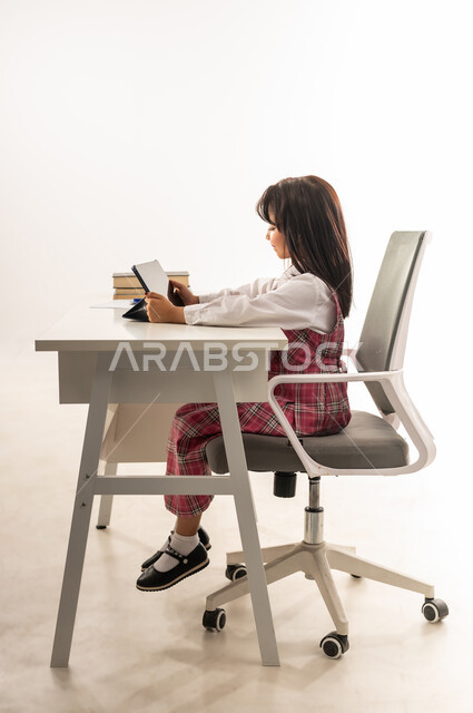 دمج التقنيات الحديثة في الحياة العلمية، مدارس و أكاديميات متطورة، طالبة سعودية تجلس على طاولة الدراسة تحمل بيدها جهاز التابلت, بورتريه من الجانب لتلميذة عربية خليجية عمانية مبتسمة ترتدي الزي المدرسي تقوم بمتابعة الدروس عن بعد عن طريق الانترنت، خلفية بيضاء