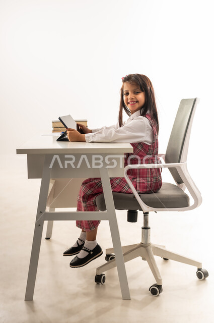 مدارس و أكاديميات متطورة، طالبة سعودية تجلس على طاولة الدراسة تحمل بيدها جهاز التابلت، دمج التقنيات الحديثة في الحياة العلمية، بورتريه من الجانب لتلميذة عربية خليجية عمانية مبتسمة ترتدي الزي المدرسي تنظر الى الكاميرا بتعابير السرور، خلفية بيضاء