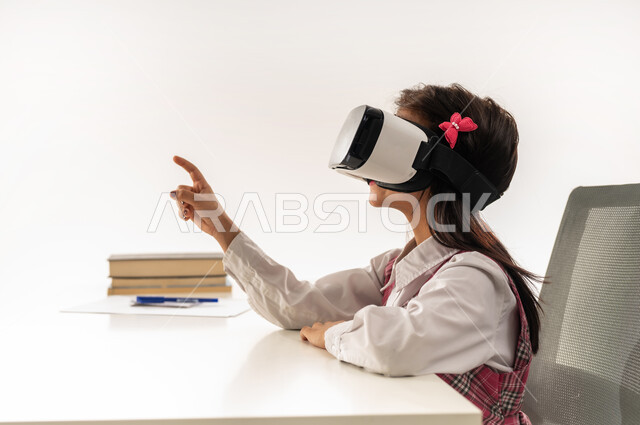 Advanced schools and academies, integration of modern technologies in scientific life, a Saudi female student sitting at a study table with books next to her, a close-up portrait from the side of an Arab Gulf Omani female student wearing a school uniform and virtual reality glasses pointing with her index finger at something, white background
