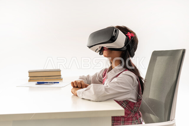اكاديميات ومدارس متطورة، دمج التقنيات الحديثة في الحياة العلمية، طالبة سعودية تجلس على طاولة الدراسة وبجانبها الكتب، بورتريه مقرب من الجانب لتلميذة عربية خليجية عمانية ترتدي الزي المدرسي تستمتع بالمشاهدة عبر نظارة الواقع الافتراضي، خلفية بيضاء