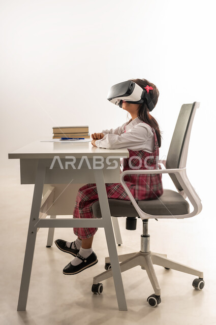 دمج التقنيات الحديثة في الحياة العلمية، طالبة سعودية تجلس على طاولة الدراسة وبجانبها الكتب، اكاديميات ومدارس متطورة، اتباع طرق تعليمية جديدة، بورتريه من الجانب لتلميذة عربية خليجية عمانية ترتدي الزي المدرسي تستمتع بالمشاهدة عبر نظارة الواقع الافتراضي، خلفية بيضاء
