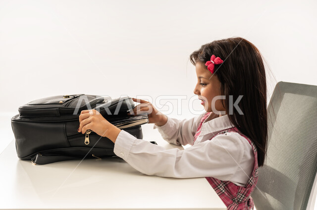 Preparing the study table to review lessons, excellence, diligence and follow-up on studies, close-up portrait from the side of an Arab Gulf Omani student wearing a school uniform pulling books from the bag, a Saudi student enthusiastic about reading, memorizing and completing the required homework, white background