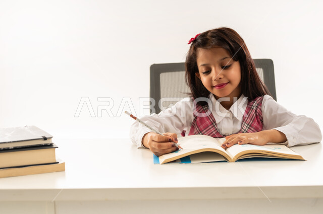 القيام بالمهام التعليمية الفعالة, اكتساب المعرفة من كتابة الواجبات، بورتريه مقرب لطالبة عربية خليجية سعودية ترتدي الزي المدرسي تجلس على طاولة الدراسة, تلميذة عمانية تقوم بتدوين المعلومات لتمكين وفهم الدروس, ايماءات الاندماج والتركيز، خلفية بيضاء