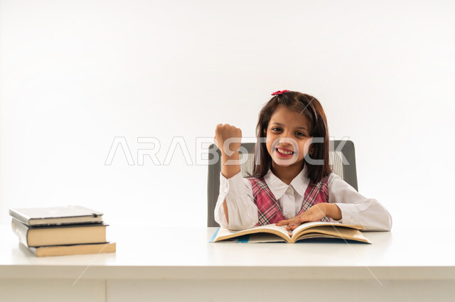 ايماءات الفرح والانتصار لمعرفة الجواب، الاجتهاد والتفوق ومتابعة الدراسة، بورتريه مقرب لتلميذة عربية خليجية عمانية ترتدي الزي المدرسي تجلس على الطاولة ترفع قبضة يدها للاعلى بحركات الفوز، طالبة سعودية تنظر الى الكاميرا بتعابير السرور و السعادة، خلفية بيضاء