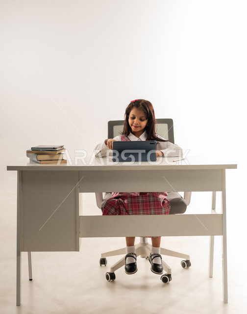 Using modern advanced technologies in scientific life, the concept of distance learning, a portrait of an Arab Gulf Omani student wearing a school uniform and next to her virtual reality glasses, a Saudi student working on a tablet looking at the camera with expressions of pleasure, white background