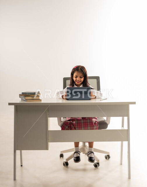 مفهوم التعلم عن بعد ,استخدام التقنيات الحديثه المتطورة في الحياة العلمية، بورتريه لطالبة عربية خليجية عمانية ترتدي الزي المدرسي و بجانبها نظارات الواقع الافتراضي، تلميذة سعودية تقوم بالعمل على جهازالتابلت تنظر الى الكاميرا بتعابير السرور، خلفية بيضاء