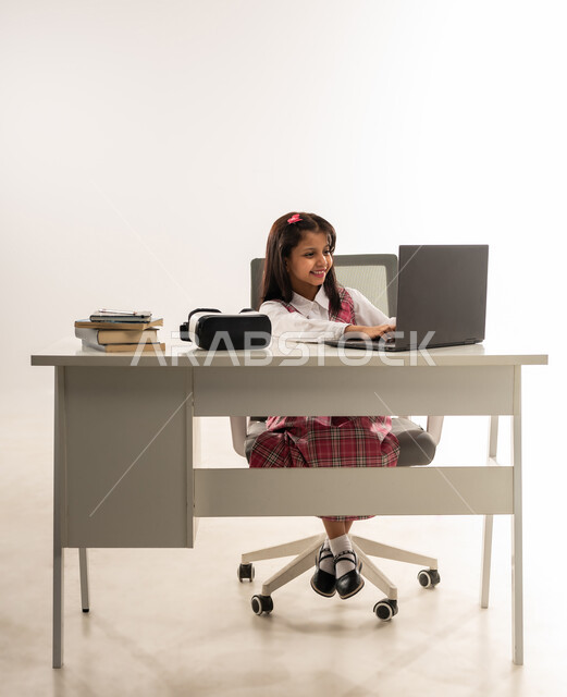 استخدام التقنيات الحديثه المتطورة في الحياة العلمية، مفهوم التعلم عن بعد ,بورتريه لطالبة عربية خليجية عمانية ترتدي الزي المدرسي و بجانبها نظارات الواقع الافتراضي، تلميذة سعودية تقوم بالعمل على جهاز الحاسوب المحمول بتعابير السرور، خلفية بيضاء