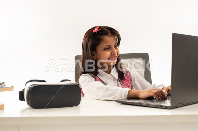 The concept of distance learning, the use of modern advanced technologies in scientific life, a close-up portrait of an Arab Gulf Omani student wearing a school uniform and next to her virtual reality glasses, a Saudi student working on a laptop with expressions of pleasure, white background