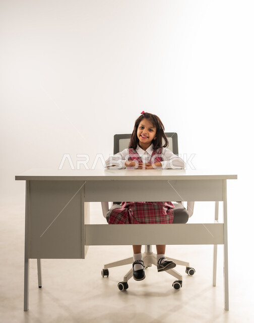Expressions of enthusiasm for returning to school, looking at the camera with gestures of happiness and pleasure, an Arab Gulf Omani student wearing a school uniform preparing to start the lesson, a portrait of a Saudi student sitting at the table with gestures of joy, white background