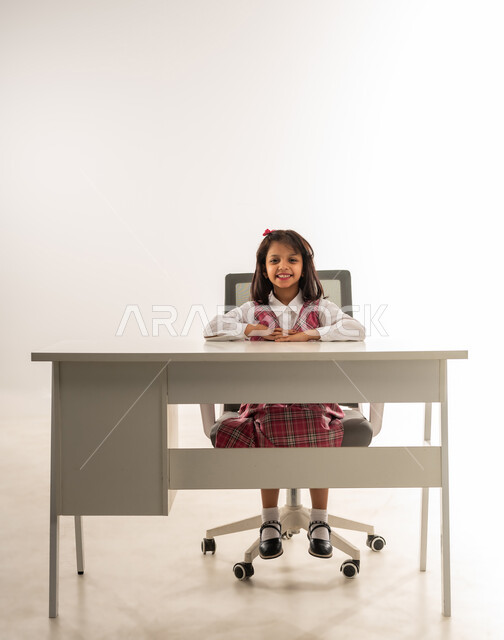 النظر الى الكاميرا بايماءات السعادة والفرح, تلميذه عربيه خليجية عمانية ترتدي الزي المدرسي تستعد لبدء الدرس, بورتريه لطالبة سعودية تجلس على الطاولة بتعابير السرور, مفهوم العودة الى المدارس, خلفية بيضاء